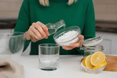 Vrouw mengt baking soda met azijn, glas water en schijfjes citroen op tafel.